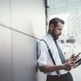 Mann mit Bart der an einer Wand anlehnt und etwas auf seinem Handy anschaut